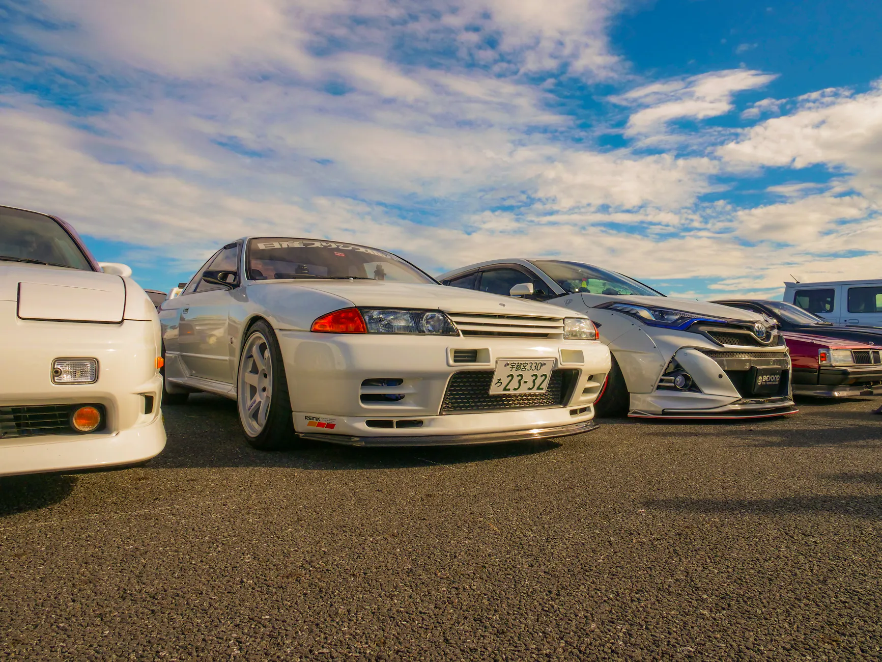 A lineup of classic JDM cars featuring a Nissan Skyline R32 at an outdoor car meet.