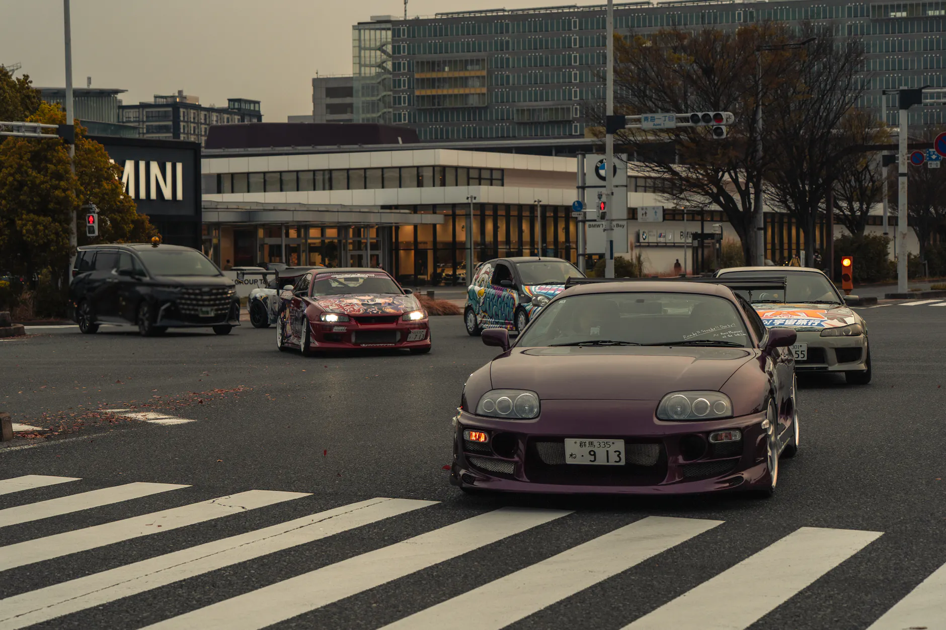 Sports cars cruising through an intersection in Tokyo, Japan, featuring modified vehicles in an urban setting.