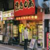 A man stands outside a Japanese restaurant in a lively street, examining the menu. Bright signage and Japanese characters create an inviting atmosphere.