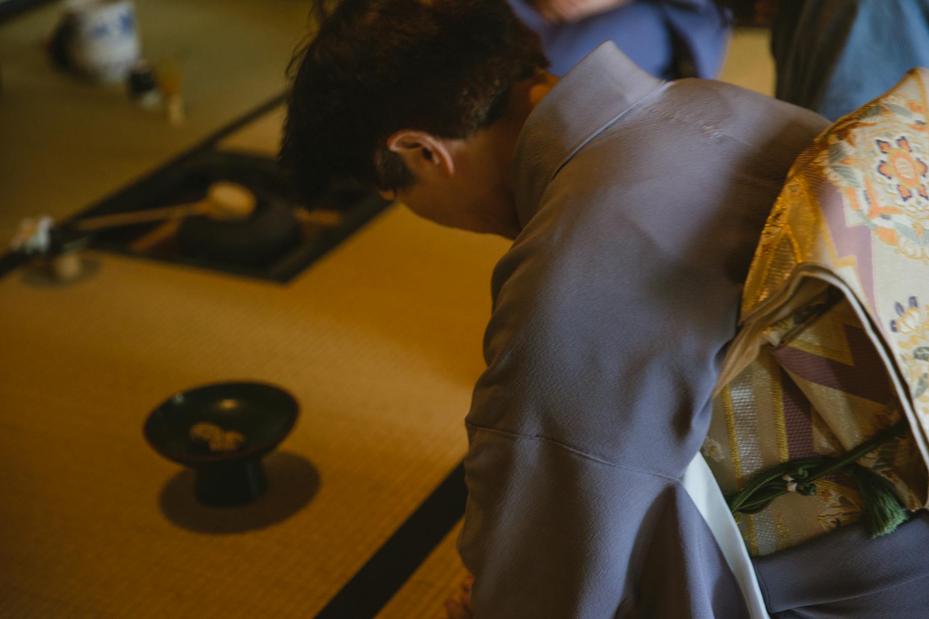 Side view of anonymous male in authentic robe bowing head while sitting on tatami with dishware for traditional tea ceremony