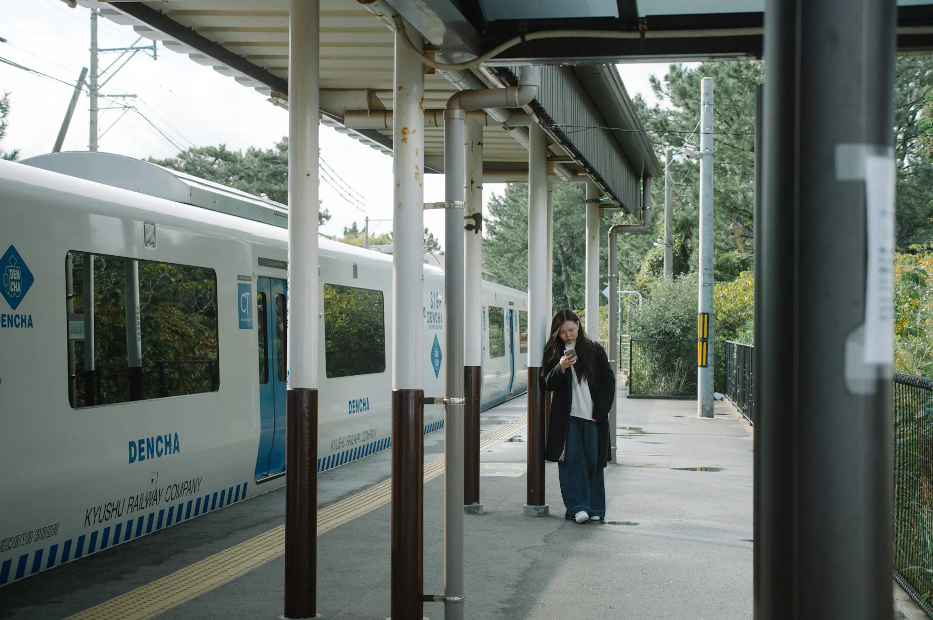 A woman checking her phone at a Japanese train station beside a Dencha train.
