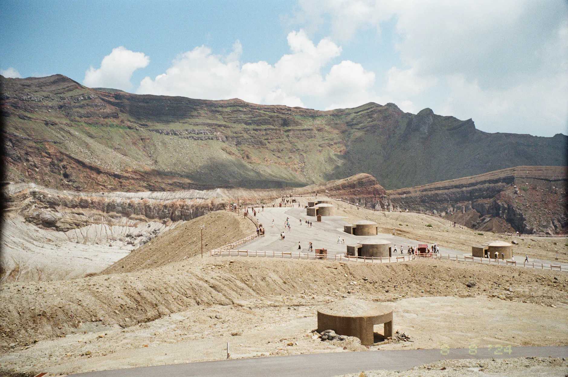 Tourists walking around the scenic crater area of Mount Aso, Japan, under a bright blue sky.