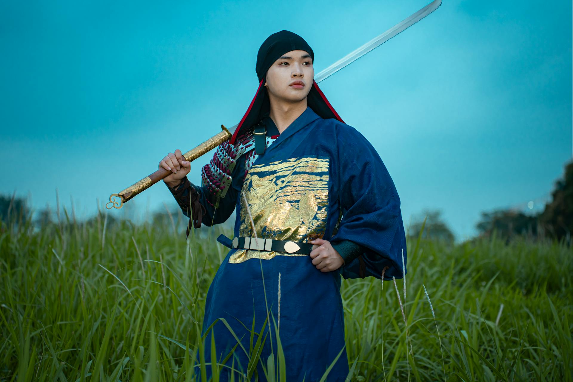 A samurai in traditional attire holds a sword in a grassy field during twilight